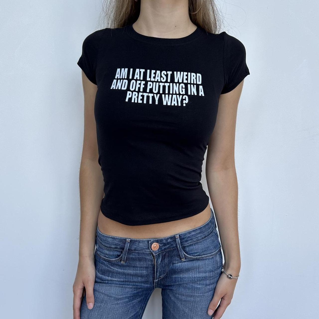 Model wearing black baby tee with "Am I At Least Weird And Off Putting in a Pretty Way" written in white across the chest.